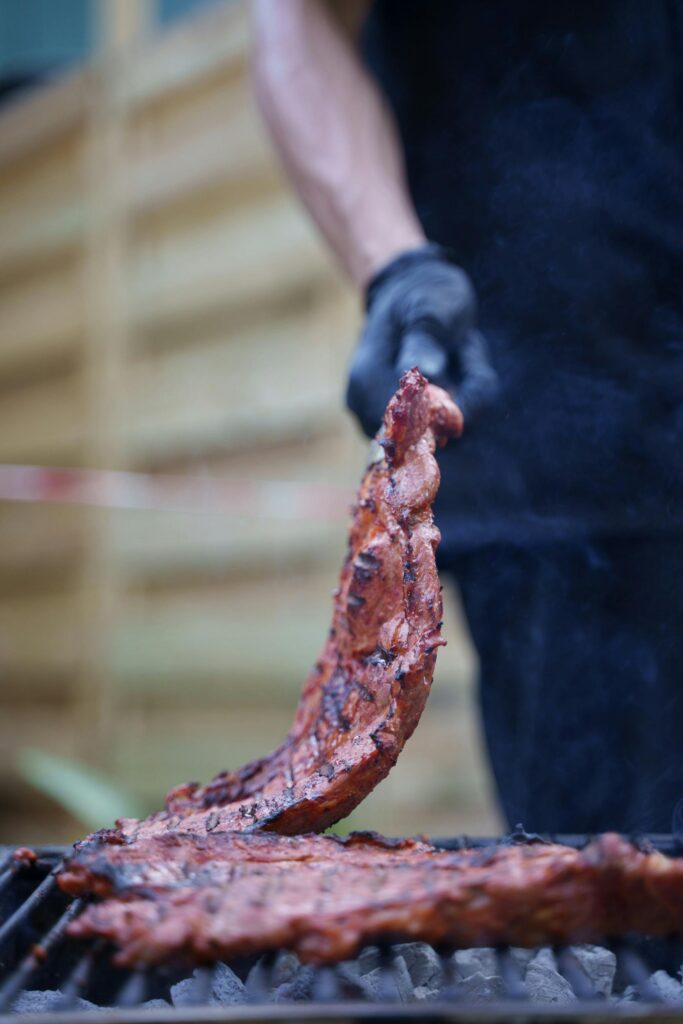 Close-up of ribs being grilled at an outdoor barbecue with a person handling meat.